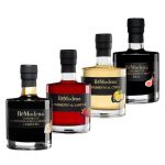 Vier Flaschen „Aceto balsamico di Modena I.G.P. 250 ml BIO“ stehen in einer Reihe, jede mit einem schwarzen Verschluss und einem einzigartigen Etikett: Auf jeder Flasche sind die Geschmacksrichtungen Trüffel, Himbeere, Zitrusfrüchte und Feige mit Abbildungen der jeweiligen Zutaten dargestellt.